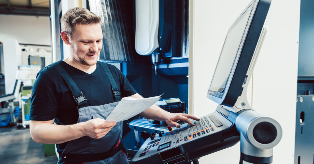 A factory worker holding a piece of paper standing in front of a computer next to a CNC machine and pressing buttons.