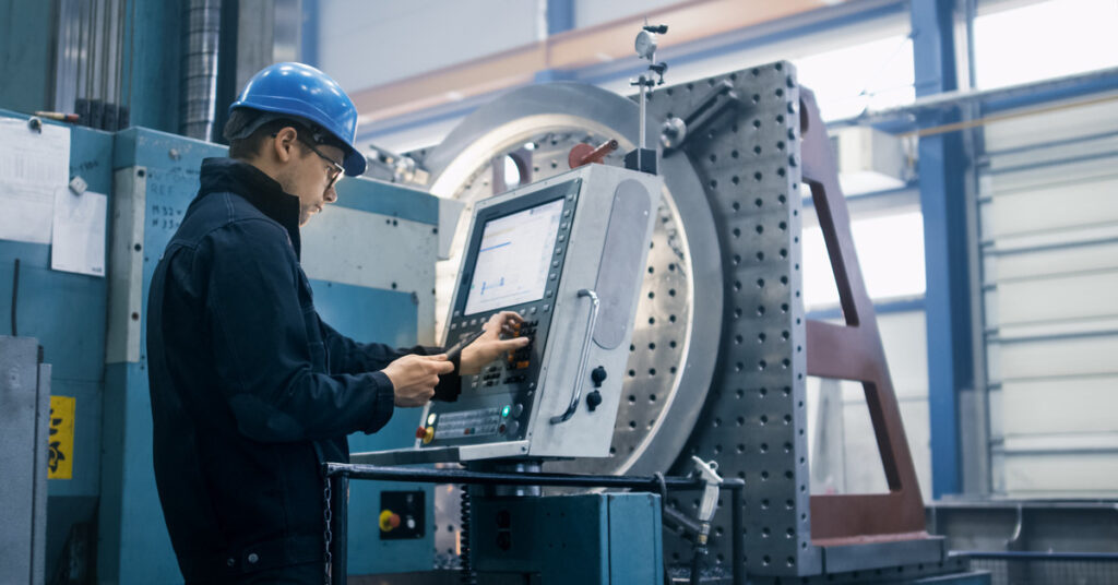 A man wearing a helmet and a uniform that is standing in front of a computer near a CNC machine and pressing buttons.